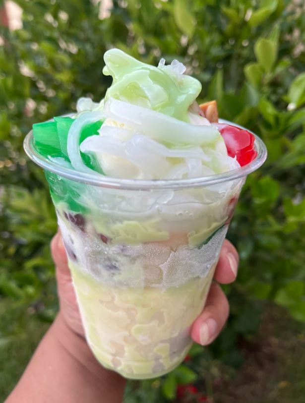 Hand holding a layered dessert drink with green, white, and red colors against a blurred green background.